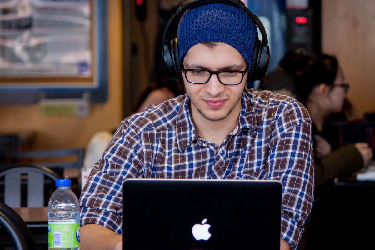 A male student is wearing a beanie, glasses and headphones while working on his laptop in a cafe.